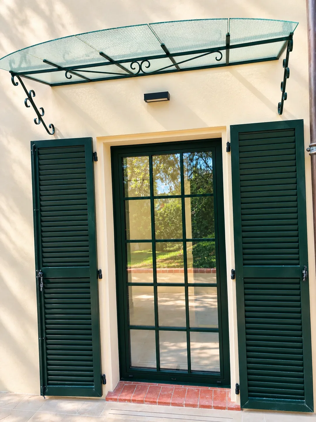 Marquise vitrée au-dessus d’une porte-fenêtre entourée de volets.